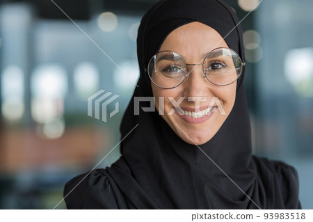 Close-up portrait of Muslim businesswoman in hijab smiling and looking at camera, female worker in glasses working inside modern office building. 93983518