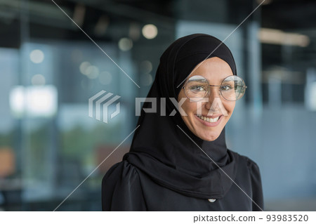 Close-up portrait of Muslim businesswoman in hijab smiling and looking at camera, female worker in glasses working inside modern office building. Close-up portrait of Muslim businesswoman in hijab smiling and looking at camera, female worker in glasses working inside modern office building. 93983520