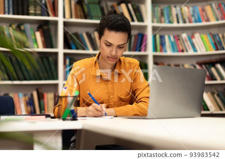 Serious woman sitting at desk in library using laptop and taking notes in notebook, studying online, copy space 93983542