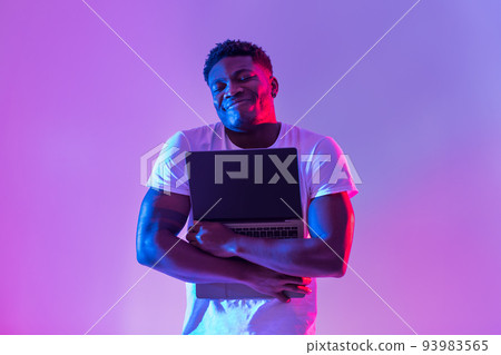 Cheerful young black man hugging laptop with empty screen in neon light, mockup for website 93983565