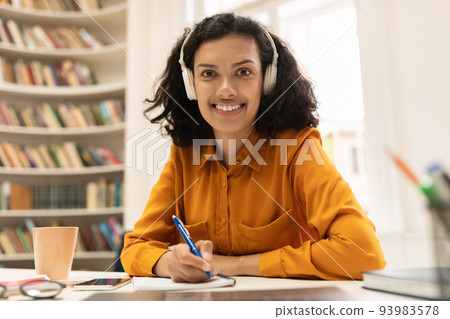 Happy woman in headset having online lesson on laptop, writing in notebook and smiling at camera, sitting in library Happy woman in headset having online lesson on laptop, writing in notebook and smiling at camera, sitting in library 93983578