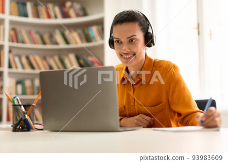 Happy female student learning online with laptop, having remote class and taking notes, sitting in library Happy female student learning online with laptop, having remote class and taking notes, sitting in library 93983609