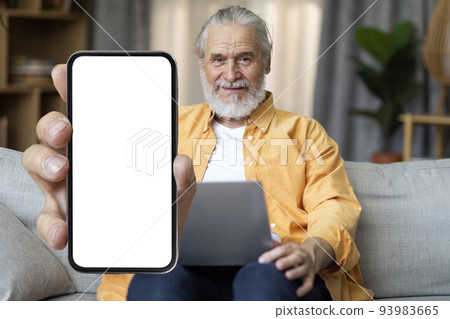 Happy elderly man sitting on couch with laptop, showing smartphone 93983665