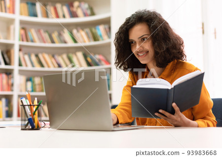 Remote education concept. Smiling mixed race lady sitting in library and using laptop, doing college home assignment 93983668