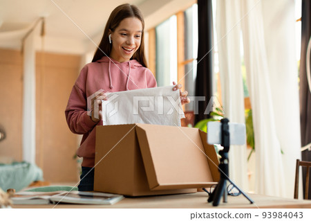 Female teen blogger shooting video, unpacking box with new clothes, talking to cellphone camera on tripod 93984043