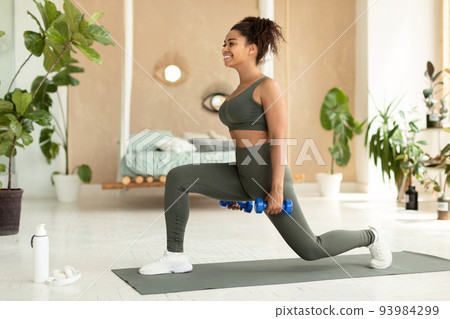 Strength training concept. Sporty african american woman doing lunges exercise with dumbbells, working out at home 93984299
