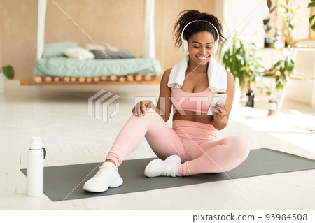 Happy black fitness lady using smartphone and wearing headphones, having break during domestic workout Happy black fitness lady using smartphone and wearing headphones, having break during domestic workout 93984508