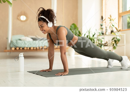 Stay at home fitness. Happy black lady doing whole body workout, standing in plank pose and listening music in headaset 93984523