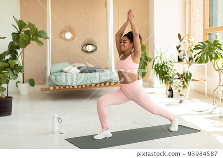 Yoga training. Fit african american lady standing in 1 warrior pose on mat, lifting hands up, training at home 93984587
