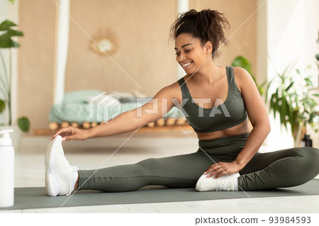 Home workout. Happy sporty black woman stretching her leg on yoga mat, warming up before training Home workout. Happy sporty black woman stretching her leg on yoga mat, warming up before training 93984593