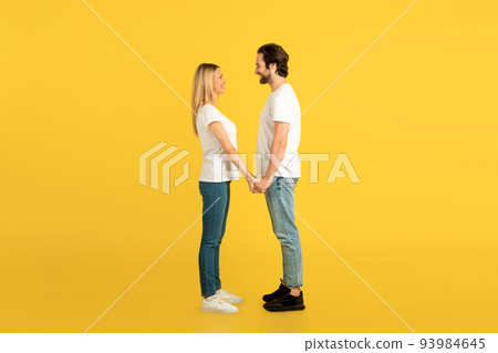 Cheerful millennial caucasian male and female in white t-shirts holding hands, looking at each other Cheerful millennial caucasian male and female in white t-shirts holding hands, looking at each other 93984645