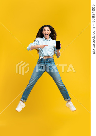 Excited lady pointing at smartphone with empty screen, jumping up in air, offering space for mobile app or website 93984809