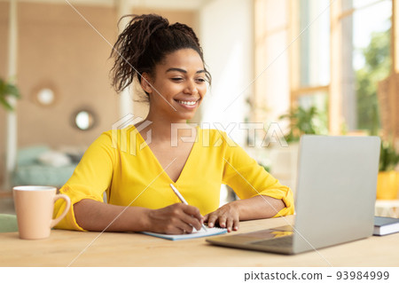 Smiling african american lady working on laptop taking notes in notebook, sitting at desk at home, copy space Smiling african american lady working on laptop taking notes in notebook, sitting at desk at home, copy space 93984999