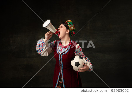 Astonished young woman in national traditional folk Ukrainian attire shouting at megaphone isolated over dark vintage background. Sport, betting 93985002