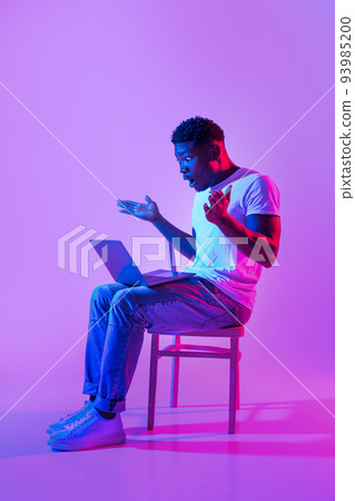 Emotional young black man sitting on chair with laptop, feeling shocked, making mistake in business project in neon light 93985200