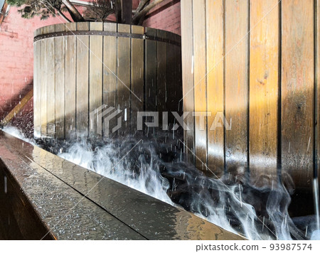 Raindrops falling on a street lamp embedded in the floor, against the background of large wooden barrels at night. Water falling on a hot lamp evaporates. 93987574