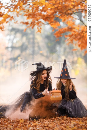 Mother witch and her daughter making a potion in autumn forest. Mother teach her daughter. Young woman and little girl wearing witches Halloween costumes and cone hats. 93989082