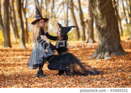 Woman with little girl walking in autumn forest. Mother play with her daughter. Mother and daughter wearing witches Halloween costumes and cone hats. Woman with little girl walking in autumn forest. Mother play with her daughter. Mother and daughter wearing witches Halloween costumes and cone hats. 93989101