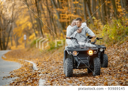 A young caucasian man and woman riding an ATV quad bike in autumn forest. Two friends maneuvering off-road ATV. Couple wearing grey sportive costumes. A young caucasian man and woman riding an ATV quad bike in autumn forest. Two friends maneuvering off-road ATV. Couple wearing grey sportive costumes. 93989145