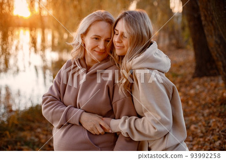 Adult daughter hugs her pregnant mom on a sunny autumn day in nature. Blonde women standing in park near the lake. Women wearing beige clothes. 93992258