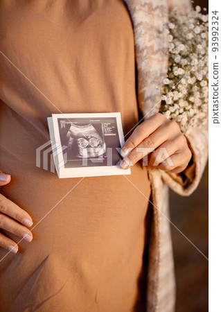 Close up of an expecting pregnant woman wearing a brown shirt and beige jacket and holding her belly outdoor. A cropped image of pregnant woman in the autumn park. Woman holding a ultrasound image of Close up of an expecting pregnant woman wearing a brown shirt and beige jacket and holding her belly outdoor. A cropped image of pregnant woman in the autumn park. Woman holding a ultrasound image of 93992324