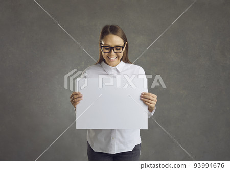Young beautiful happy woman stands on a gray background and holds a blank white sheet in his hands. 93994676