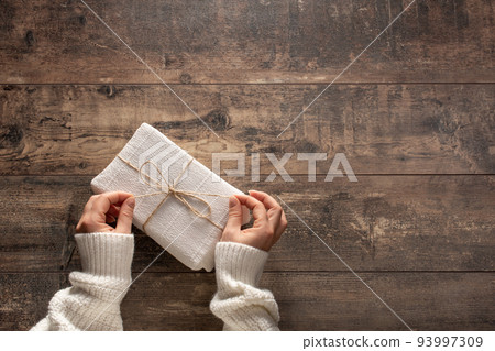 Gift box concept. Hands in sweater holding christmas gift. Christmas gift. Congratulations.Top view of woman holding traditional decorated gift box. Hands holding gift box on wooden background. 93997309