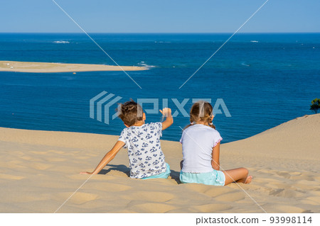 Girl and boy sitting on a huge sand dune, famous tourist destination Dune Pyla. Pilat Dune in France. Girl and boy sitting on a huge sand dune, famous tourist destination Dune Pyla. Pilat Dune in France. 93998114