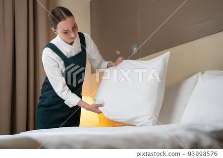 Female worker makes bed changing linen and folding pillows Female worker makes bed changing linen and folding pillows 93998576
