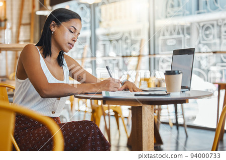 Student writing, taking notes or doing a project assignment in cafe or restaurant using laptop and notebook. Young woman taking an online education class or doing study work, learning in coffee shop. 94000733