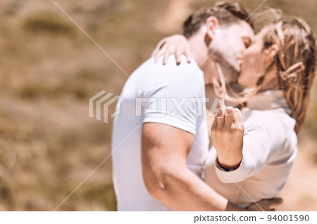Young woman showing her engagement ring finger while couple kiss enjoying a sweet moment together. In love, romantic and kissing couple ready for marriage loving romance in the sun and nature 94001590