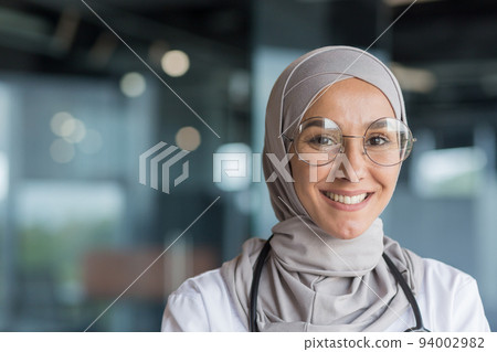 Close-up photo portrait of Muslim female doctor in gray hijab, female doctor smiling and looking at camera, female worker in glasses and white medical coat and glasses working inside modern clinic 94002982
