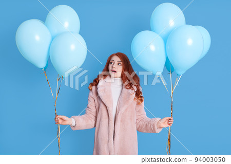 Shocked worried young ginger woman girl wearing fur coat, celebrating, posing on pastel color background with air balloons, keeping mouth opened, looking aside, looks astonished and shocked. 94003050