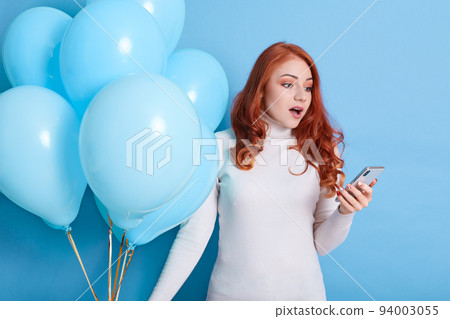 Shocked young woman in white sweater and jeans posing isolated over blue background, celebrating birthday, holds air balloons using mobile phone, keeps mouth widely opened, expresses astonishment. Shocked young woman in white sweater and jeans posing isolated over blue background, celebrating birthday, holds air balloons using mobile phone, keeps mouth widely opened, expresses astonishment. 94003055