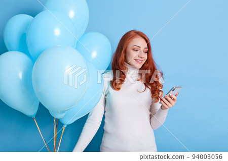 Satisfied beautiful European birthday girl has pleasant texting conversation, receives congratulation, wears white shirt, holds party balloons, celebrates holiday. 94003056