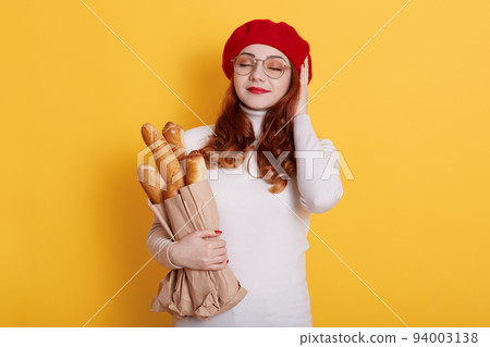 Pretty young redhead woman stands with closed eyes and pleasant smile. Glad ginger girl feels happy isolated on yellow wall, holding long bread baguettes, wearing red beret and white shirt. Pretty young redhead woman stands with closed eyes and pleasant smile. Glad ginger girl feels happy isolated on yellow wall, holding long bread baguettes, wearing red beret and white shirt. 94003138