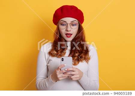 Image of shocked student girl in red beret posing with cellphone in hands against yellow background, looks at device's screen with opened mouth, wears red lips, expressing shock. 94003189
