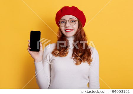 Image of smiling red haired girl wearing red beret and white shirt, girl with red lips holding smart phone with blank screen in hands, isolated on yellow. Copy space or promotion text. 94003191