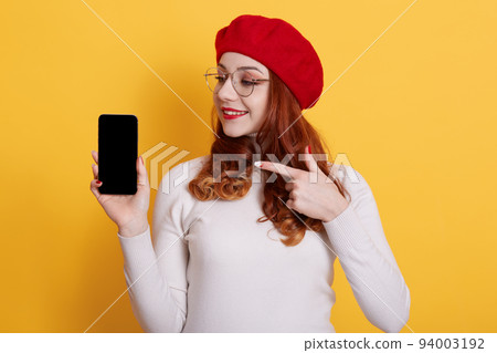 Smiling happy red haired female holding mobile phone with blank screen, pointing at device with fore finger, girl curls standing isolated over yellow background, copy space for promo. Smiling happy red haired female holding mobile phone with blank screen, pointing at device with fore finger, girl curls standing isolated over yellow background, copy space for promo. 94003192