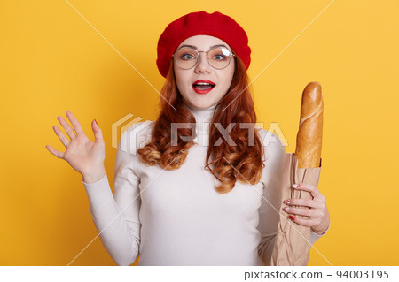 Portrait of beautiful astonished young woman holding paper bag with bread baguette against yellow background, lady with red hair with surprised expression, raises hands and keeps mouth opened. Portrait of beautiful astonished young woman holding paper bag with bread baguette against yellow background, lady with red hair with surprised expression, raises hands and keeps mouth opened. 94003195