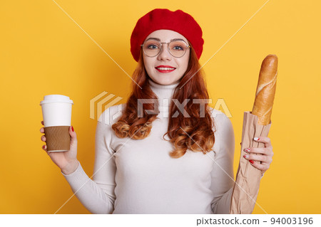 Portrait of girl with long red hair wearing beret, white shirt and glasses, looks at camera and smiles, holding bread in paper bag and take away coffee, model posing against yellow wall. Portrait of girl with long red hair wearing beret, white shirt and glasses, looks at camera and smiles, holding bread in paper bag and take away coffee, model posing against yellow wall. 94003196