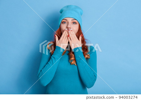 Stunned red haired woman with wavy hair, covers both hands on mouth, afraids of something,looks astonished, dressed in casual shirt, isolated over blue background, stands speechless indoor. 94003274