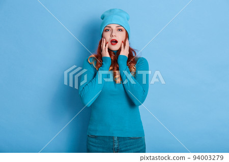 Ginger girl looks at camera with widely opened mouth, touches her cheeks with palms, lady wearing casual attire and cap, posing against blue wall, woman wears red lipstick. Ginger girl looks at camera with widely opened mouth, touches her cheeks with palms, lady wearing casual attire and cap, posing against blue wall, woman wears red lipstick. 94003279