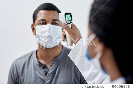 Male covid patient getting his temperature taken with medical equipment by a doctor in hospital consultation room. Man wearing mask and health care professional pointing digital infrared thermometer. 94006868