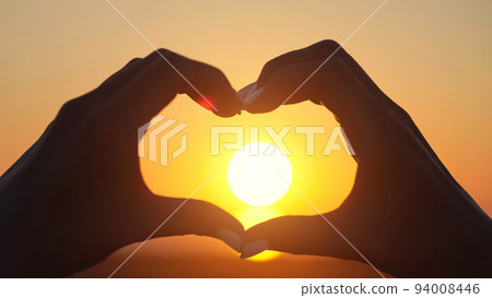 Woman makes big heart with hands at back sunset closeup Woman makes big heart with hands at back sunset closeup 94008446