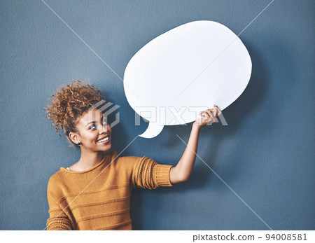 Young woman holding a speech bubble against a grey background in a studio. Portrait of a casual, happy and smiling female holding copy space empty shape for social media, chat and voice or message. Young woman holding a speech bubble against a grey background in a studio. Portrait of a casual, happy and smiling female holding copy space empty shape for social media, chat and voice or message. 94008581