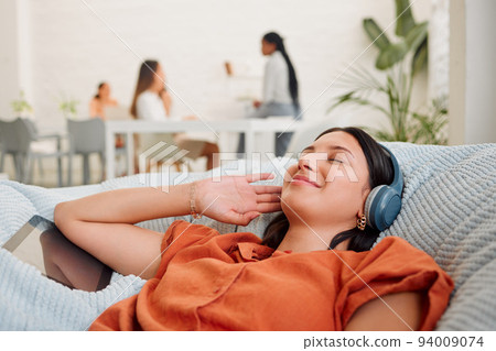 Creative wearing headphones, listening to music from tablet and relaxing, feeling inspired or motivated in a casual workspace. Happy and zen business woman lying on office bean bag and taking a break 94009074