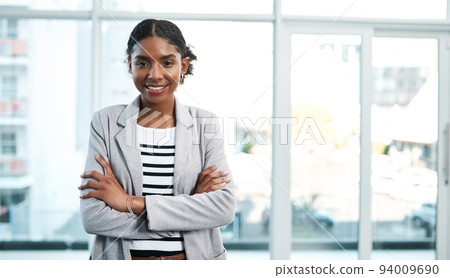 Business woman standing with arms crossed, looking proud and confident in an office alone at work. Portrait of a young black female manager, boss or employee smiling, looking successful and powerful Business woman standing with arms crossed, looking proud and confident in an office alone at work. Portrait of a young black female manager, boss or employee smiling, looking successful and powerful 94009690