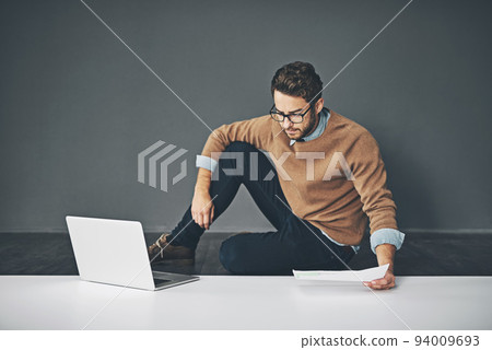 Business man working on a laptop, reading a report and going through finance paperwork while sitting on the floor in an office at work. Serious professional manager looking at papers with copyspace 94009693