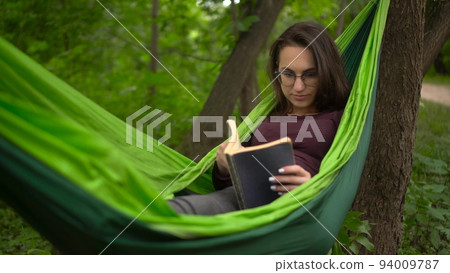 A young woman lies in a hammock and reads a book. A girl with glasses in nature is engaged in self-development. 94009787